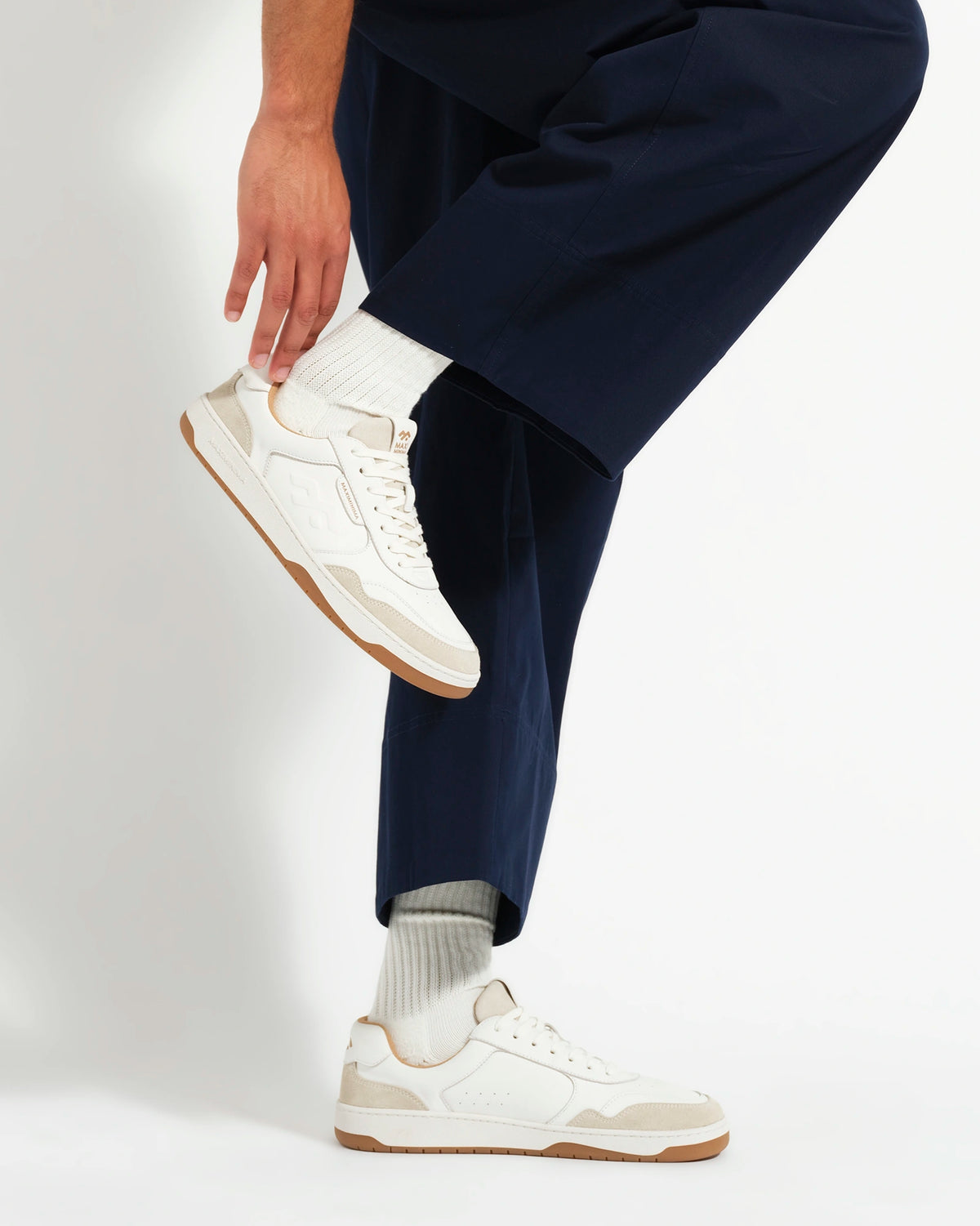A person wearing white low-top barefoot sneakers with a gum sole, paired with white socks and navy blue trousers, shown from the waist down against a light gray studio background.